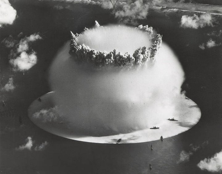 763px-Crossroads_baker_explosion.jpg - Crossroads Baker, showing the white surface "crack" under the ships, and the top of the hollow spray column protruding through the hemispherical Wilson cloud. Bikini Island beach in the background.